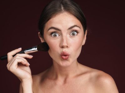 Image of excited half-naked woman making kiss lips and using makeup brush isolated over dark red background